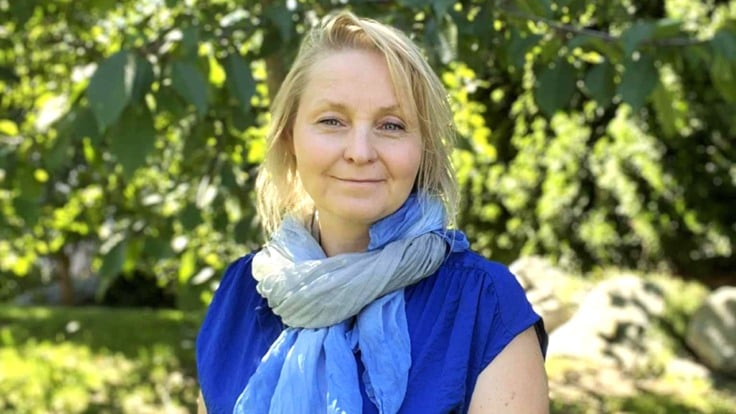 En kvinna i blå klänning står på gräsmatta, framför grönskande träd.