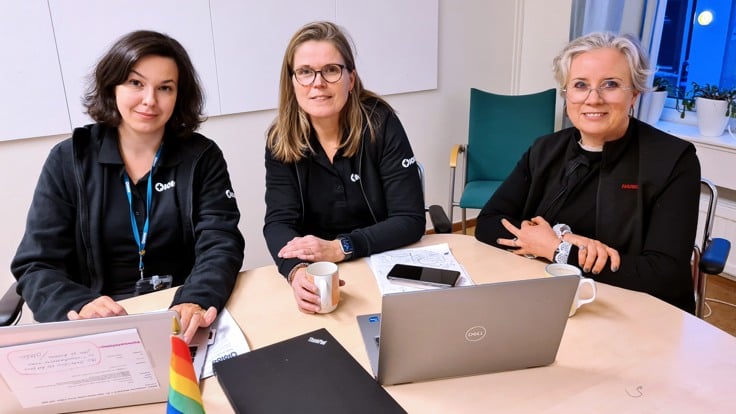 Tre kvinnor sitter vid ett konferensbord. Framför sig har de en dator. På bordet står en liten regnbågsfärgad flagga.