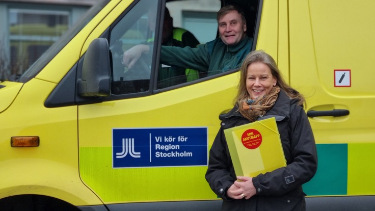 Kvinna står med en gul mapp vid sidan av en ambulans. Ambulansföraren och kvinnan tittar i kameran.
