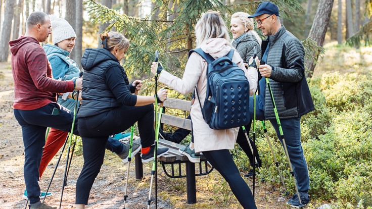 En grupp personer med gåstavar står i skog. De har sitt ena ben på en bänk.