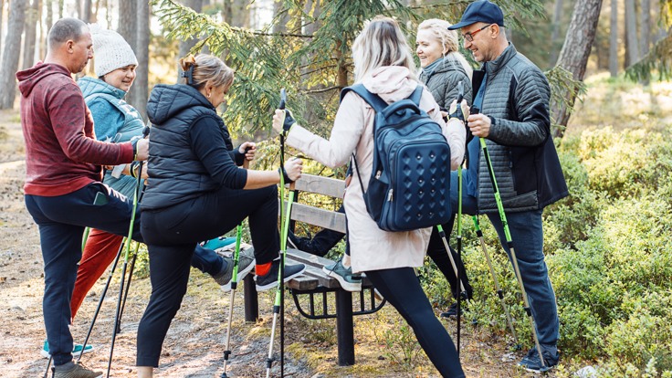 En grupp personer med gåstavar står i skog. De har sitt ena ben på en bänk.