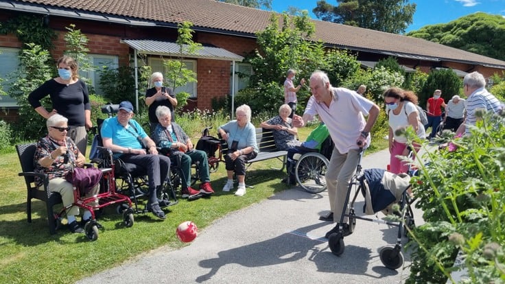 Äldre man rullar iväg ett bowlingklot ute i solen, många äldre tittar på.