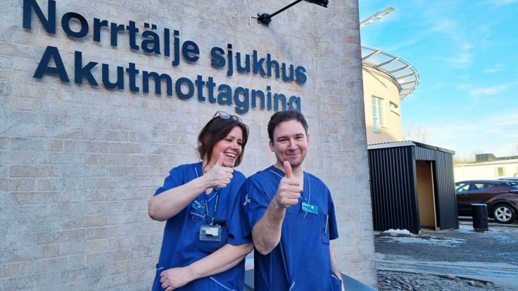 En man och en kvinna i blå sjukhuskläder skrattar och gör tummen upp utanför akutmottagningen på Norrtälje sjukhus.