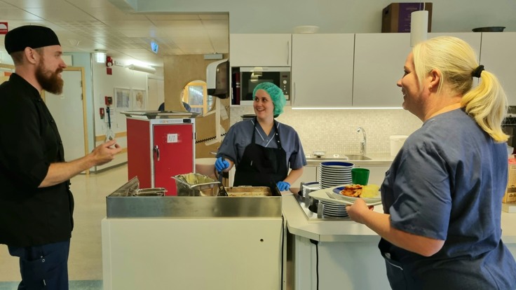 Man och två kvinnor klädda i blå vårdkläder står i köksmiljö.