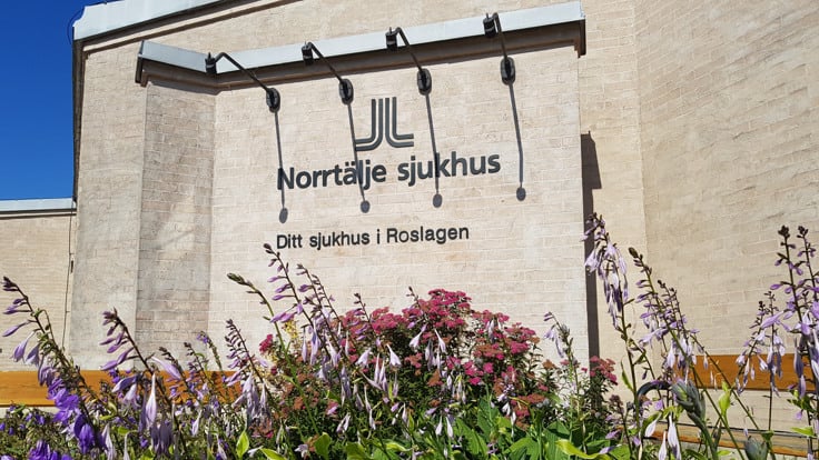 Fasad på tegelbyggnad med skylt där det står Norrtälje sjukhus och blommor i förgrunden.