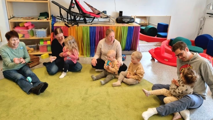 Vuxna och barn sitter på en matta på golvet i förskolemiljö.