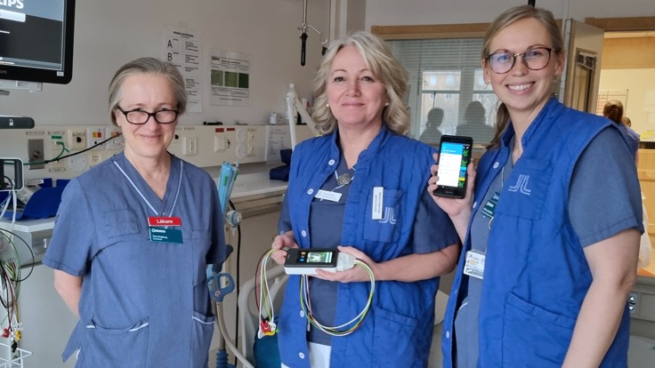 Tre kvinnor i blå bussaronger och vita byxor i ett patientrum på sjukhus.