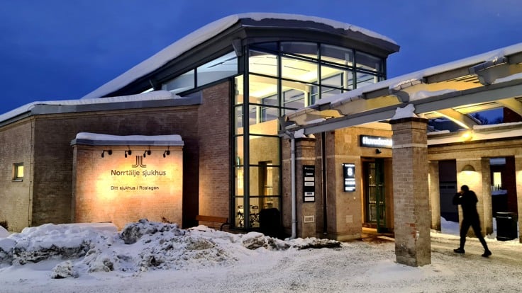 Tegelbyggnad med stort glasparti. Skymningsljus och snö på marken och taket. En person är på väg mot byggnaden. På fasaden sitter en skylt med texten "Norrtälje sjukhus".